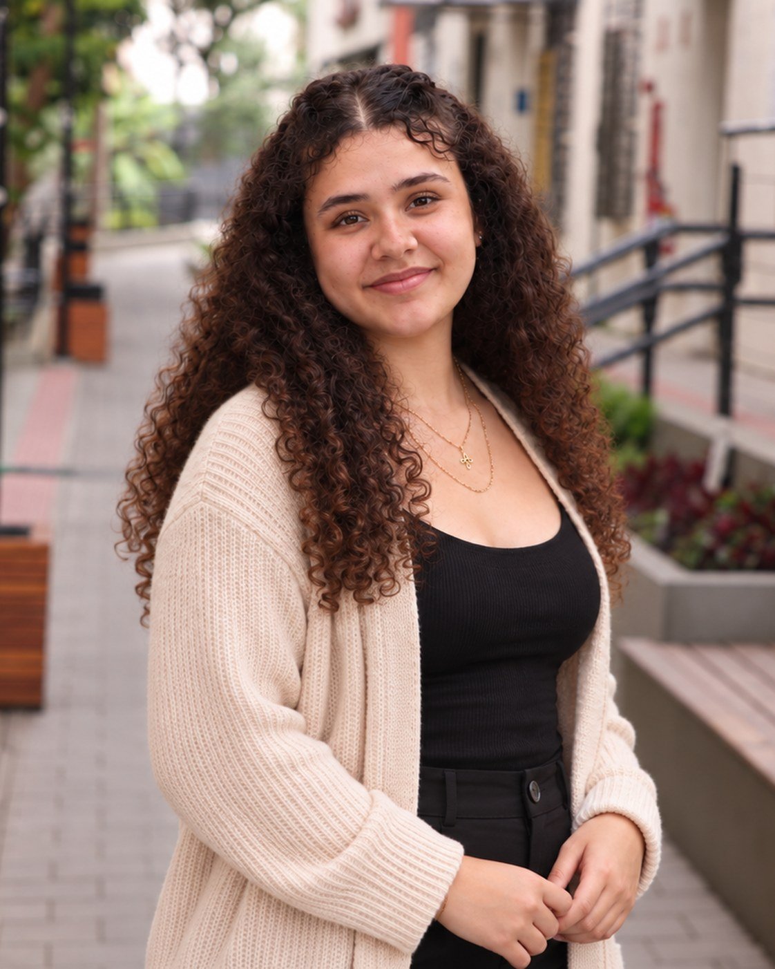 Julia Bodden, psicóloga, sorrindo de leve em uma rua arborizada, de cardigan bege